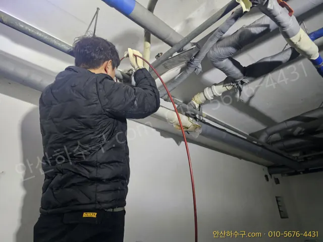 천장 횡주관 점검구에 고압 노즐 호스를 밀어 넣는 노란 조끼 작업자 뒷모습 — 아파트 공용배관 고압세척